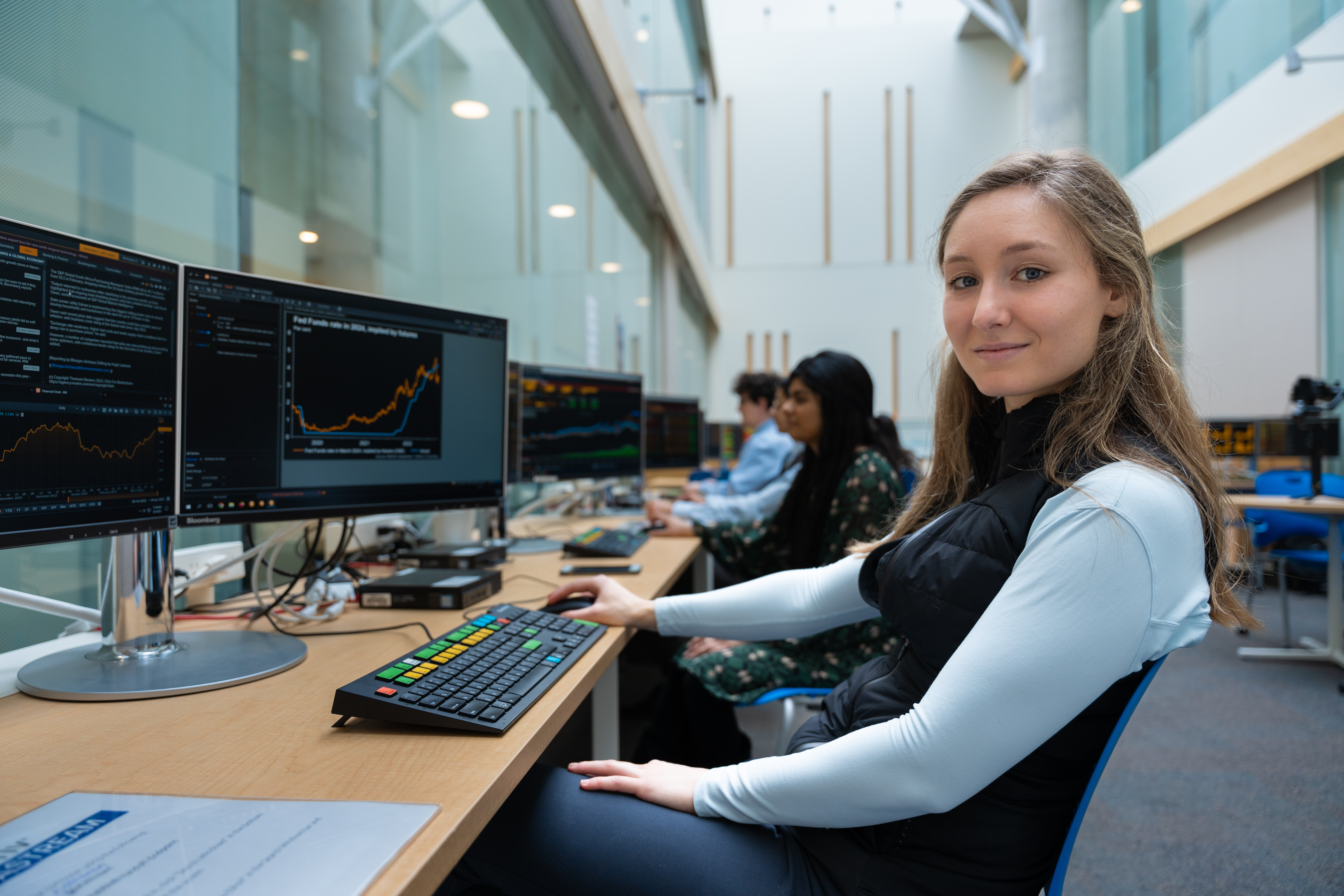 a person sitting looking at the camera. Their hand is on the mouse and there is a graph on the computer monitor