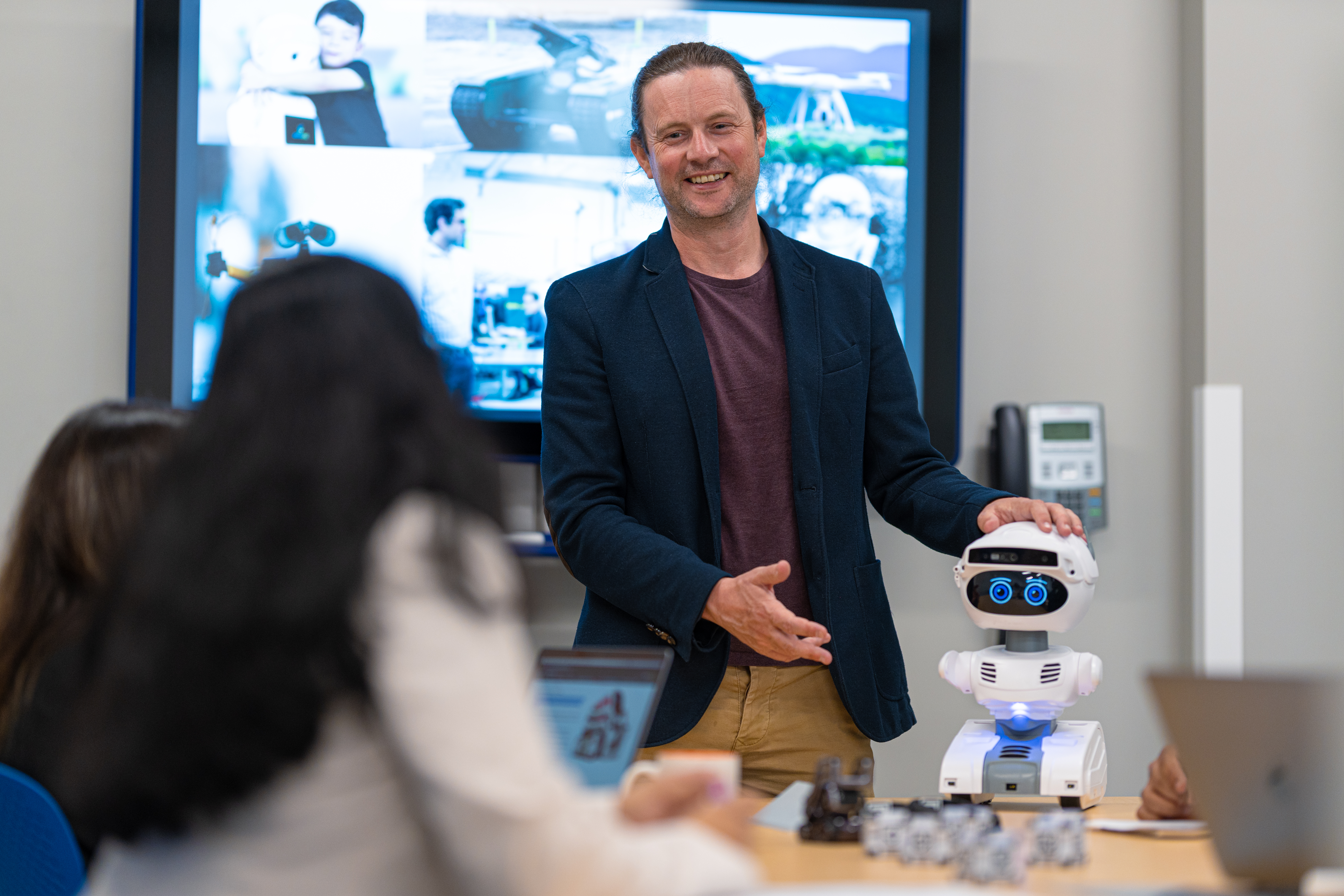 a person standing facing a group of people at a table smiling with a small robot on the table