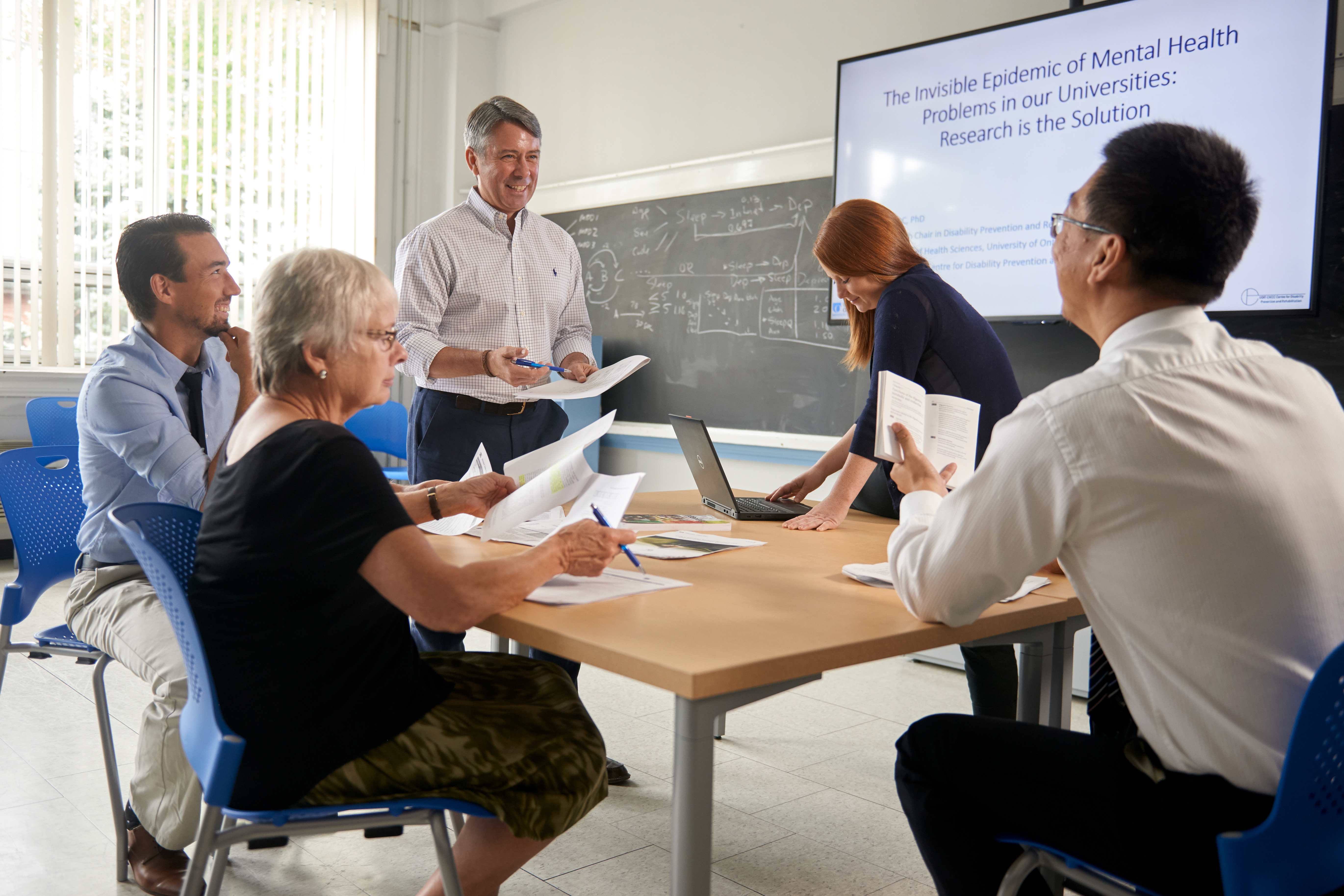 a group of people at a table sitting having a conversation. One person is looking at their laptop, one person is hold a pen and paper and one person is holding a note book. There is a projector with a PowerPoint 