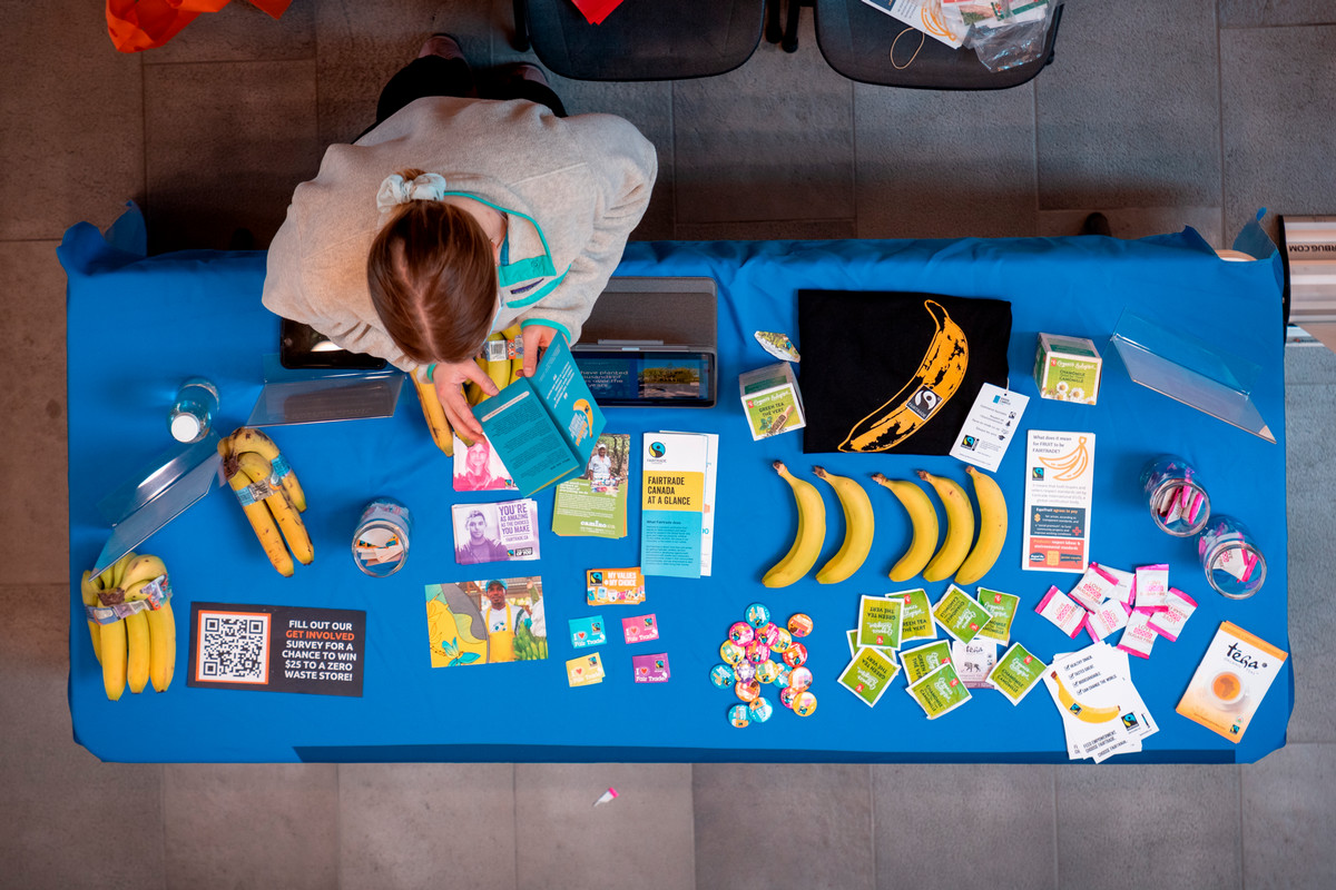 a photo of a table booth with a blue table cloth that has assorted items on it. Items included bananas, pins, flyers and tea bag packets.