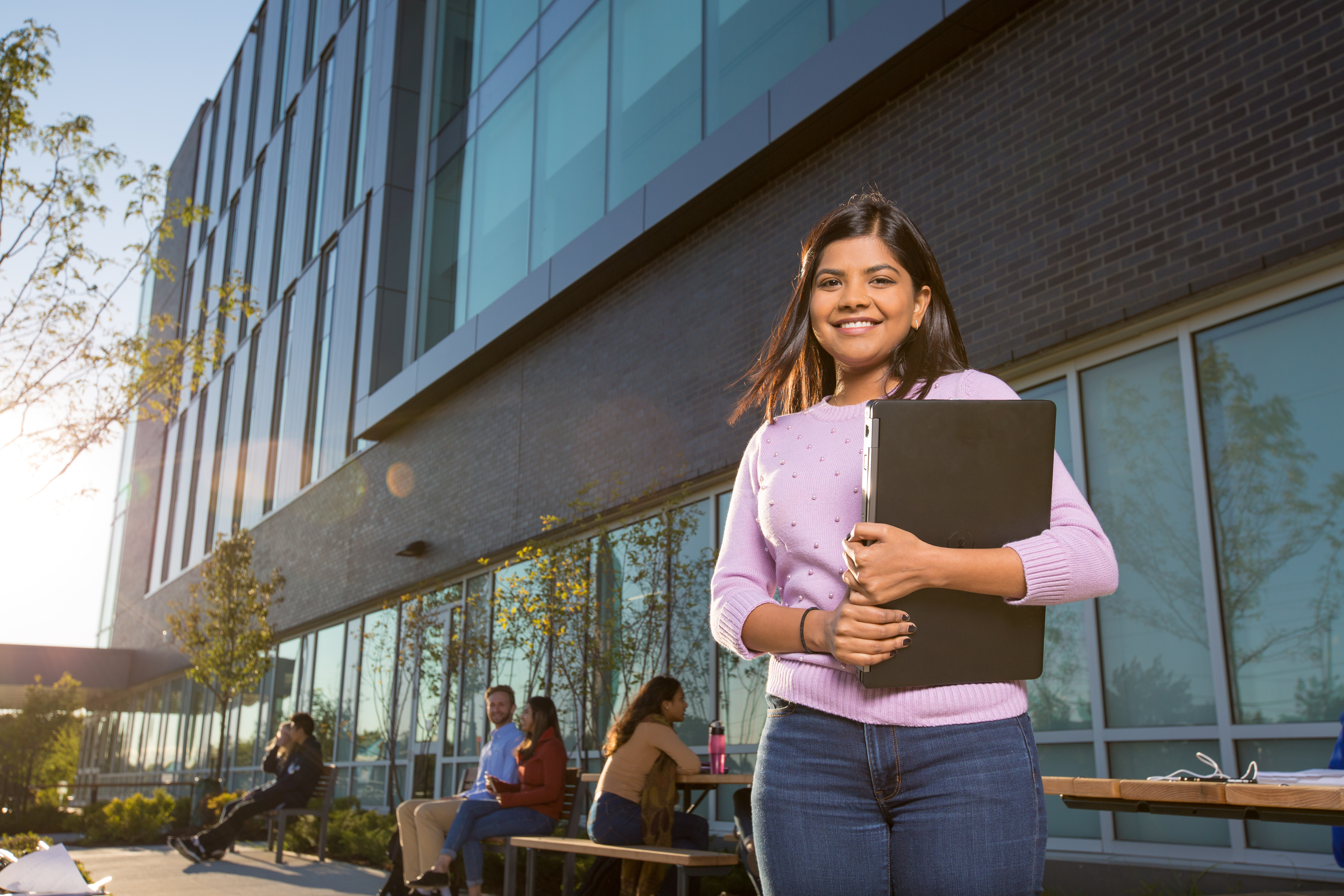 a student standing in front of the University building hold their laptop smiling