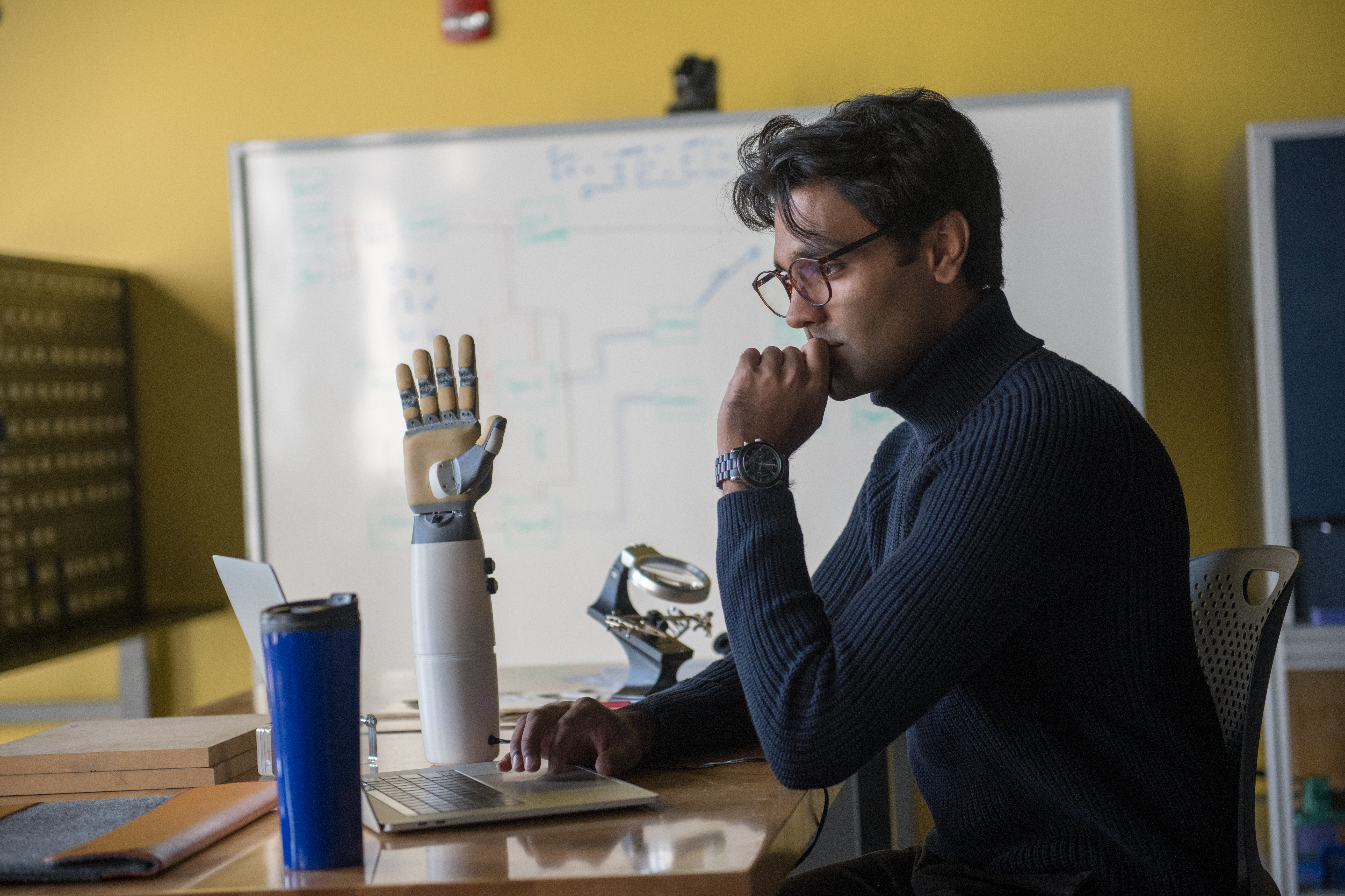 A student sitting at a table in a classroom facing their laptop with their hand over their chin.